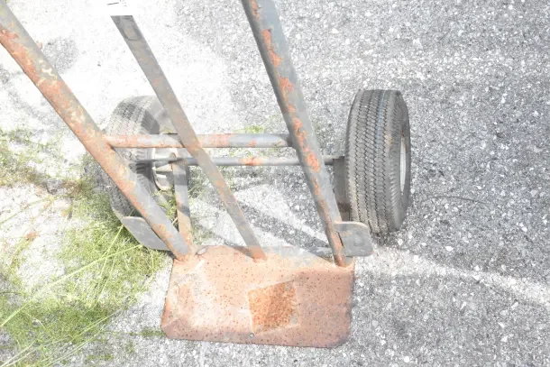 Rusty metal furniture dolly with large rubber wheels on asphalt surface. Visible wear and rust on frame.