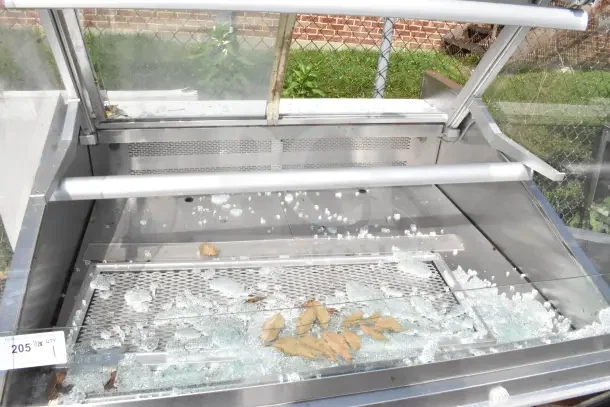 Commercial display case with broken glass, stainless steel frame, shelving, and scattered debris. Auction tag visible.