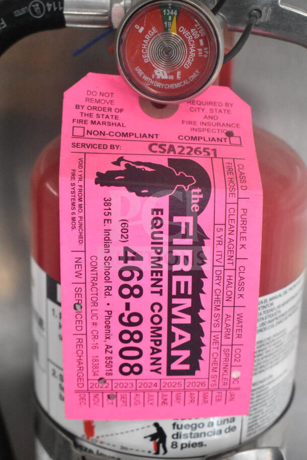 Pink safety tag on fire suppression system cylinder, marked "Compliant," serviced by CSA22651, Fireman Equipment Company.