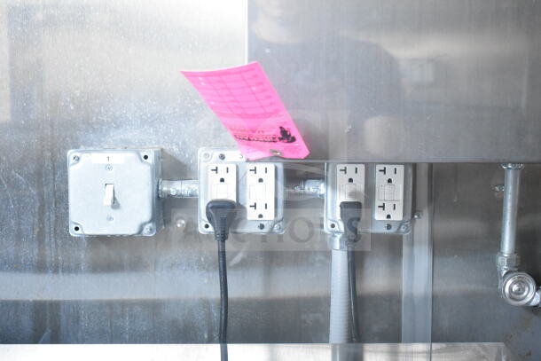 Electrical outlets with multiple plugs in use, attached to a stainless steel interior wall of a Rock Solid Cargo food trailer. A bright pink inspection tag is partially visible.