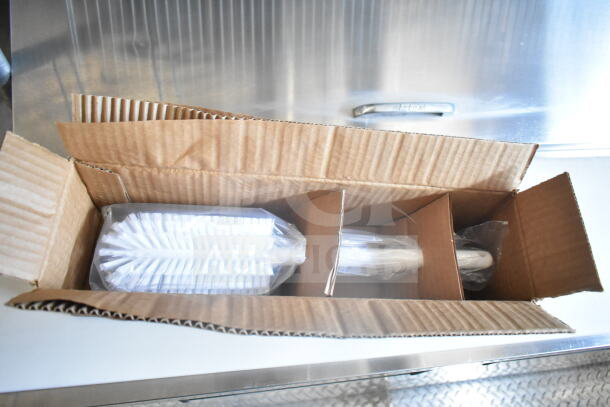 Open cardboard box containing packaged cleaning brushes on a stainless steel surface inside a food trailer.