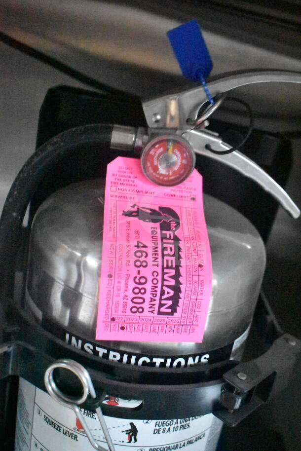 Fire extinguisher with gauge and a pink service tag from The Fireman Equipment Company, showing compliance dates.