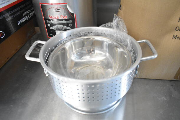 Aluminum stock pot with perforated basket and handles on a metal surface. Nearby box has restaurant kitchen labels.