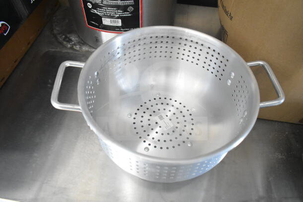 Stainless steel colander with side handles, placed on a metal surface. Background shows a stock pot with a label.