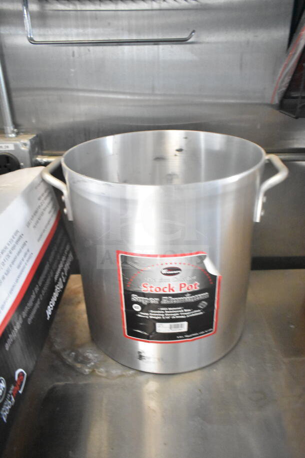 Large aluminum stock pot with dual handles, black and red label indicating "Stock Pot Super Aluminum." Appears on a stainless steel surface.
