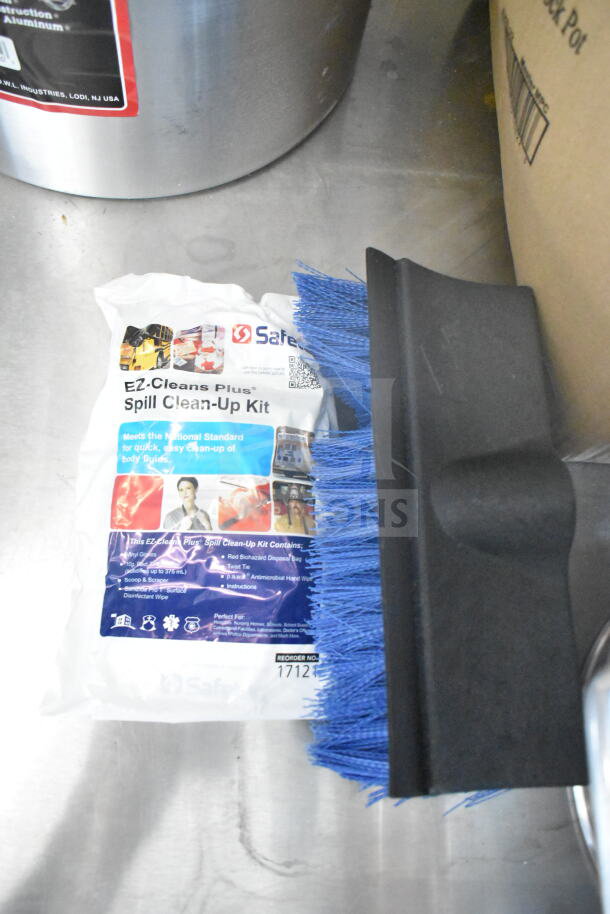 Pack of Safe-T Spill Clean-Up Kit on a metal surface with a blue brush. Includes gloves and cleaning materials.