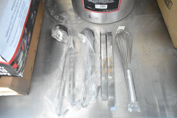 Kitchen utensils in packaging on stainless steel work surface, including ladles, tongs, and a whisk.