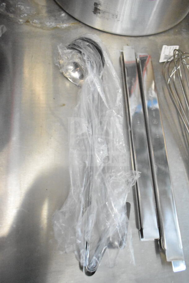Stainless steel utensils on a metal surface, including a whisk, tongs, and a sealed ladle.