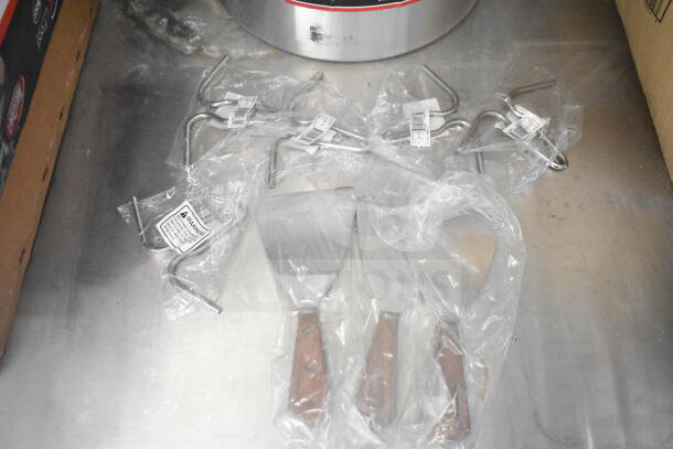 Cooking utensils on a stainless steel surface, including spatulas with wooden handles and metal hooks in plastic packaging. Labels show warnings.