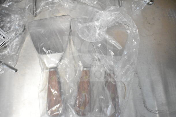 Two stainless steel spatulas with wooden handles in clear plastic packaging, set on a metal surface.