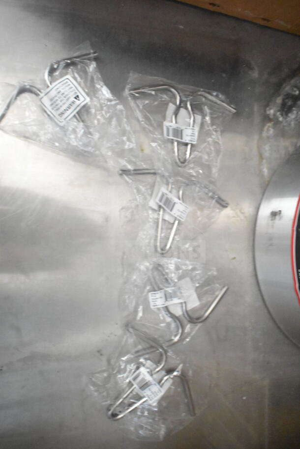 Plastic-wrapped metal hooks lying on a metallic surface, each with a label showing a barcode and product information.
