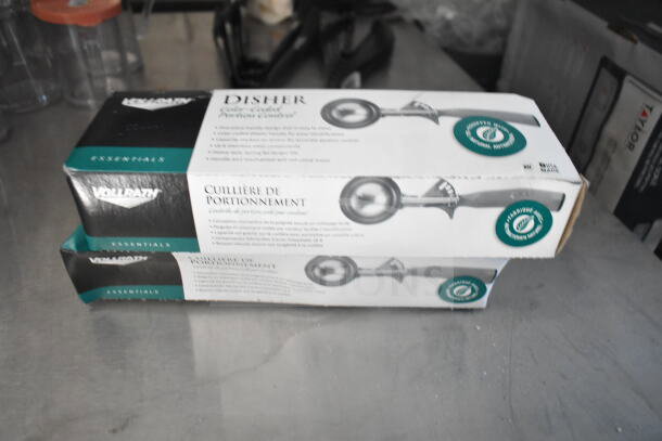 Two Vollrath Essentials disher boxes, unopened, on a countertop. Each box displays a metal scoop with ergonomic handle.