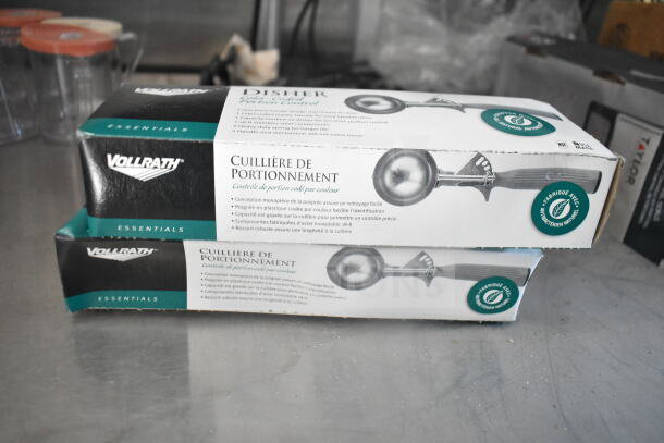 Two boxes of Vollrath Essentials disher scoops, shown on a metal surface. Packaging features color-coded portion control information.