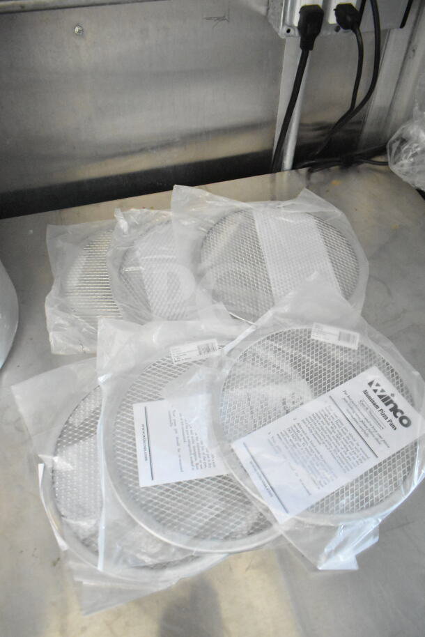 Five Winco aluminum pizza screens in packaging, placed on a stainless steel surface.