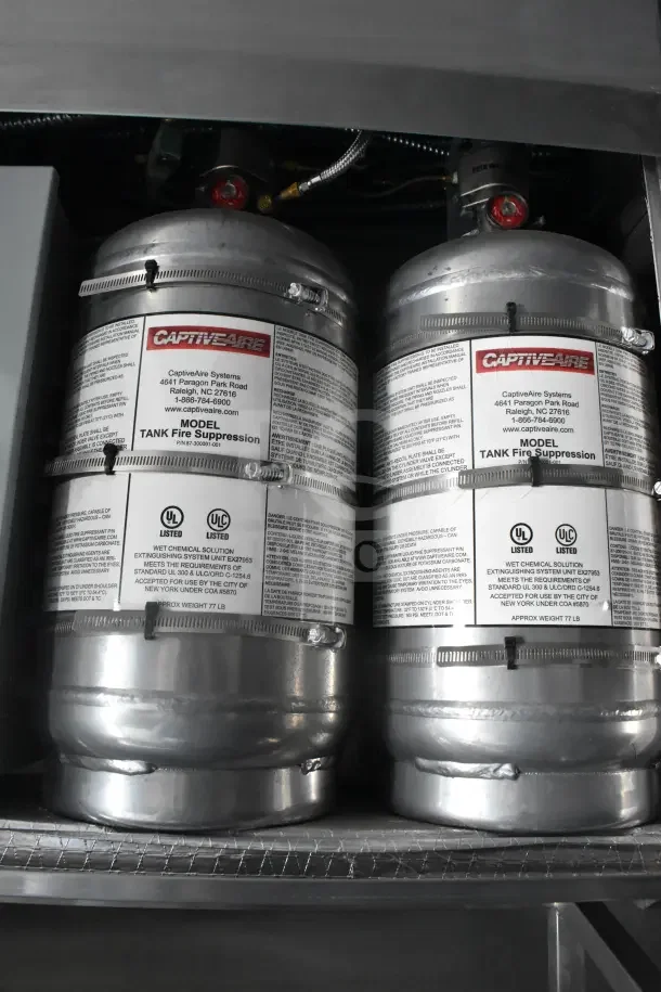Twin CaptiveAire fire suppression tanks in Texino food trailer, labeled with safety compliance details, silver metal finish.
