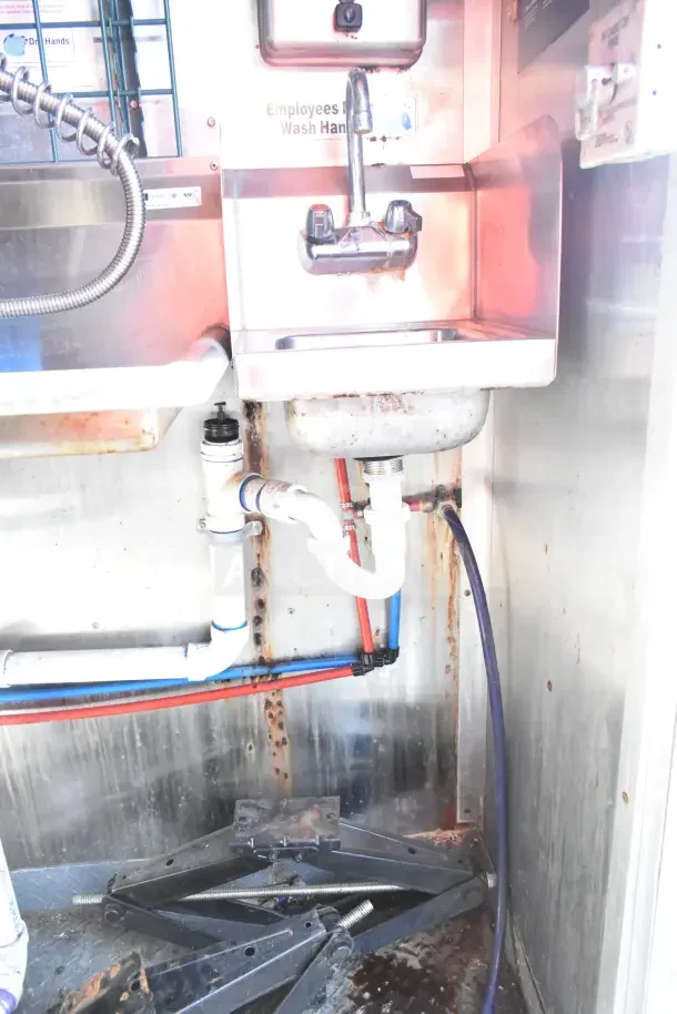 2020 Diamond Cargo kitchen trailer sink with visible plumbing and splash guard. Rust and wear on pipes and floor. "Employees Wash Hands" sign above.