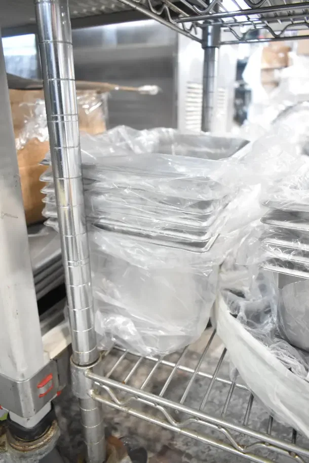 Stack of 17 new kitchen items, including 176STP1800BK black non-skid trays and stainless steel bins, wrapped in plastic.