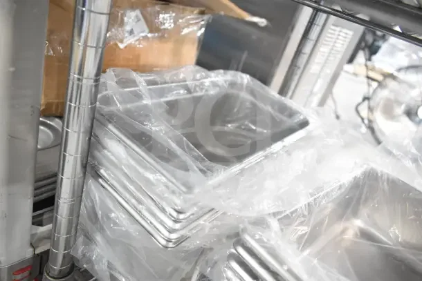 Stack of stainless steel drop-in bins, wrapped in plastic, on metal shelving. Excellent condition, bulk auction lot.