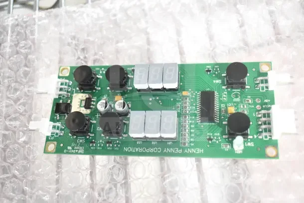 Electronics circuit board labeled "Henny Penny Corporation," set on bubble wrap, featuring capacitors, displays, and connectors.