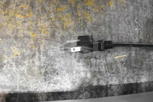 Black power plug of a commercial meat slicer's cord, placed on a scratched, yellow-streaked surface.