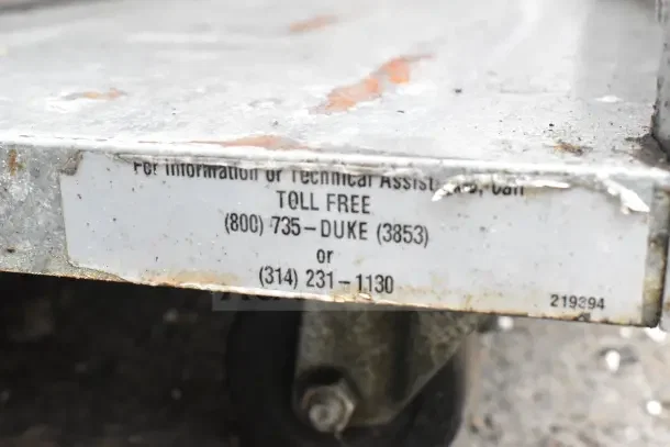 Cart with over shelf on commercial casters, metal structure, visible wear, label for technical assistance, phone numbers.
