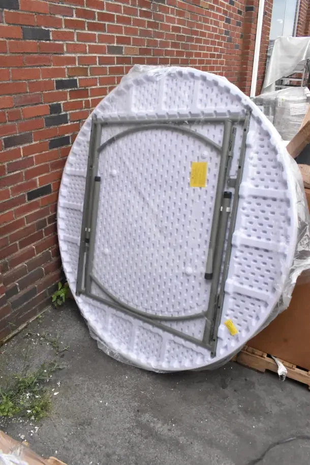 White round folding table, brand new with scratches and dents. Metal legs, plastic top, labels attached, stored upright.