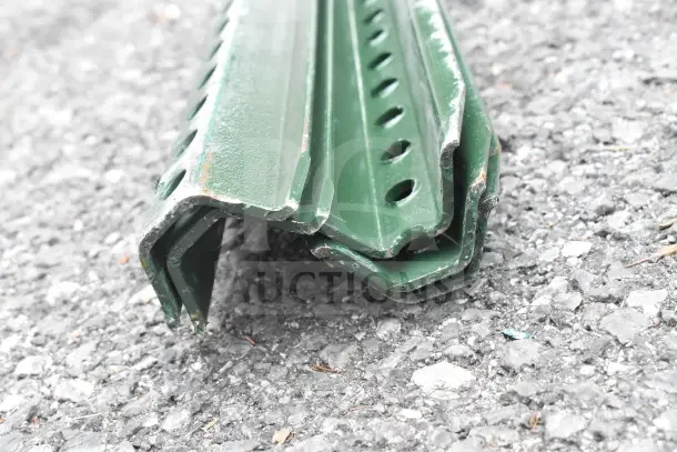 Lot of 5 green metal posts, slightly rusted edges, perforated design, stacked on pavement.