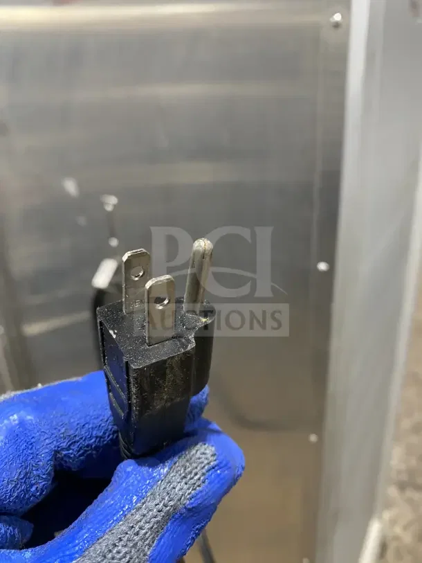 Three-prong plug of Bevles HPC6836 warming/proofing cabinet, shown gloved and held close-up, stainless steel background.