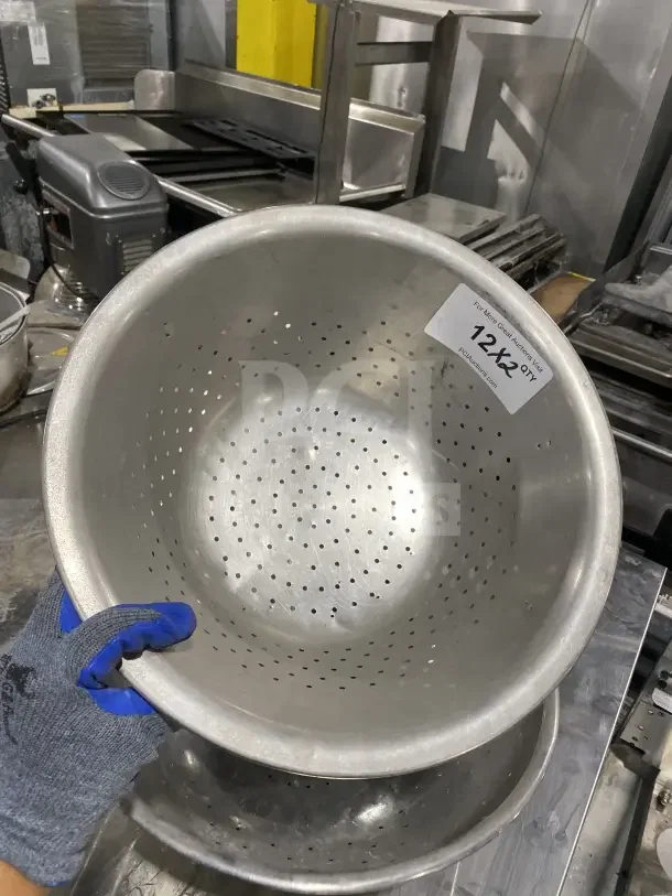 Commercial stainless steel colander, perforated design, tag reads "12X2 QTY," held by gloved hand, excellent condition.