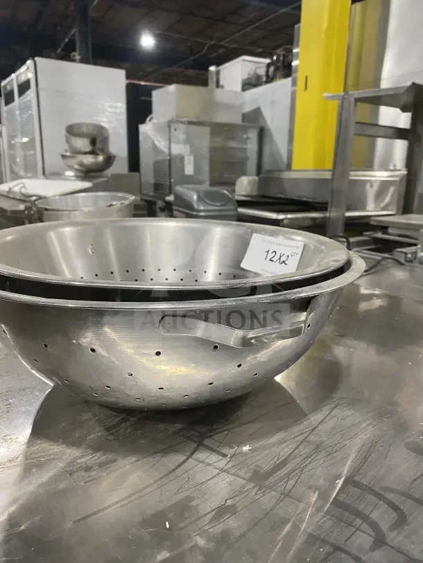 Commercial stainless steel colanders with dual handles, lightly scratched, auction tag "12x2 qty" visible.