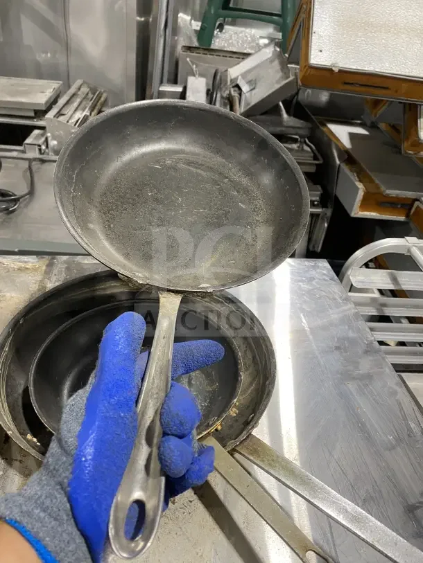 Commercial stainless steel non-stick frying pans, heavy-duty handle, signs of use, in an industrial kitchen setting.