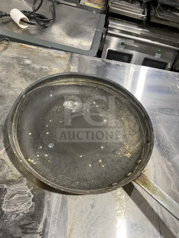Commercial stainless steel non-stick frying pan with heavy-duty handle, visibly worn with surface marks, auction item.
