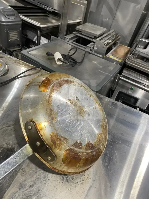 Commercial stainless steel non-stick frying pan with heavy-duty handle, visible wear and discoloration on base, used condition.