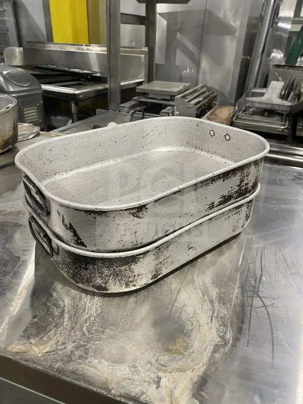 Commercial aluminum roasting and baking pans with handles, visibly worn, set of two; industrial kitchen backdrop.
