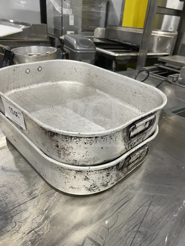 Stack of commercial aluminum roasting pans with handles, showing noticeable wear. Each pan includes a side label indicating quantity.