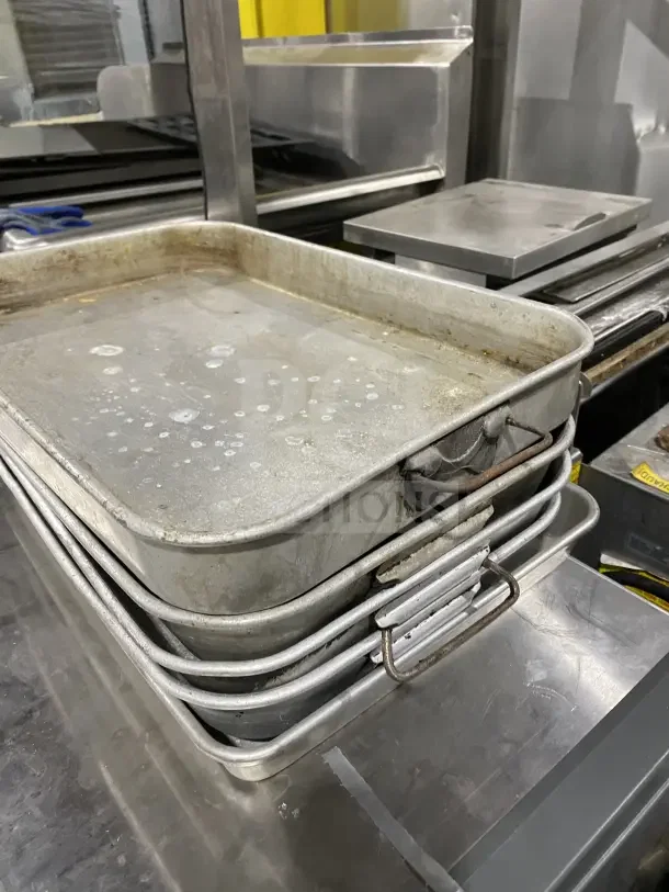 Commercial stainless steel baking trays stacked, showing signs of wear. Durable with side handles. Ideal for heavy-duty use.