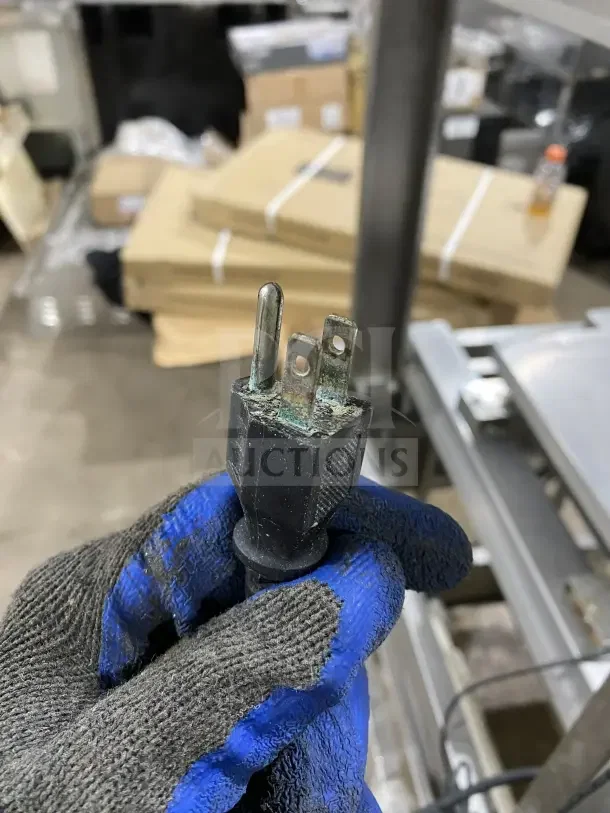 Gloved hand holding a power plug with visible corrosion; background shows part of a stainless steel food packaging station on casters by Good Nature.