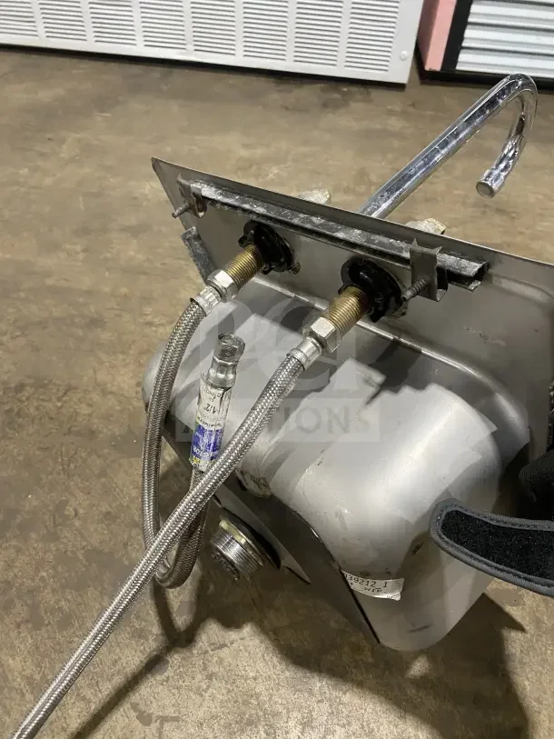 Commercial stainless steel drop-in hand sink with attached faucet, visible plumbing connections, and minor wear.