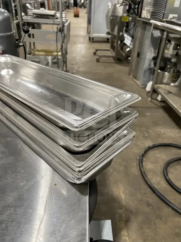 Stack of commercial stainless steel full-size hotel pans, lightly used, in a kitchen setting. Auction lot includes six pans.