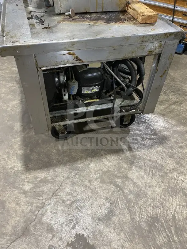 Commercial stainless steel 2-drawer refrigerated chef base on casters, heavy-duty, visible wear and surface rust, with exposed refrigeration mechanism.