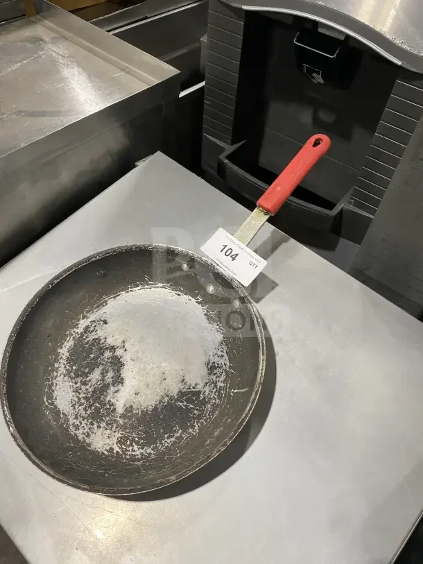Commercial aluminum non-stick fry pan with wear, featuring a removable red silicone sleeve handle, marked "104".