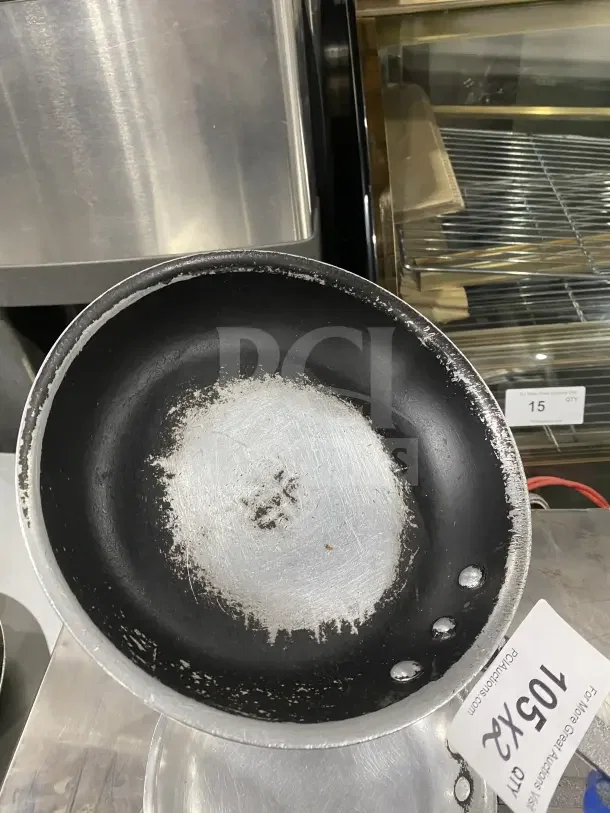 Commercial aluminum non-stick fry pan with handle, shows wear. Tag indicates auction item "105 XX". Two pans included.