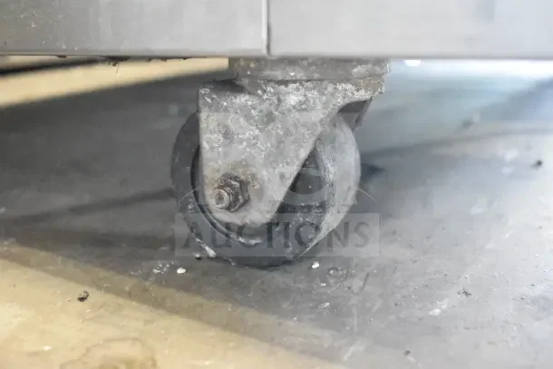 A metal commercial caster attached to the base of a kitchen appliance, showing wear and dirt.