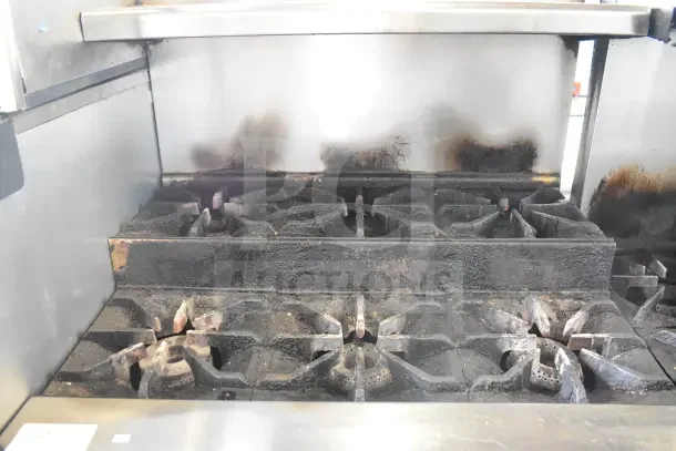 Commercial gas-powered range with 6 burners, 2 tiers, over shelf, back splash, and under shelf. Shows wear and discoloration.
