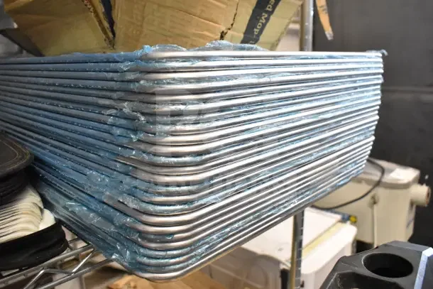 Stack of 24 brand new full-size scratch and dent baking sheets in protective wrapping on a shelf.