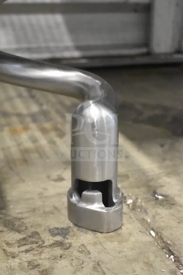 Shiny, stainless steel dough hook for mixer, brand new with visible scratches and dents, on a workshop floor.