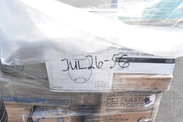 Stacked boxes labeled "JUL26-56" with various brand new scratch and dent items, including cups and bags, wrapped in plastic.