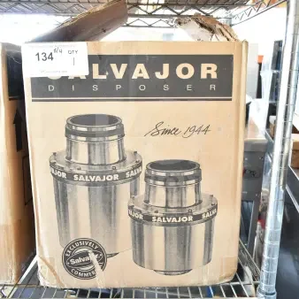 Cardboard box of Salvajor disposer, marked "Since 1944," on metal shelving. Auction label "134" visible.