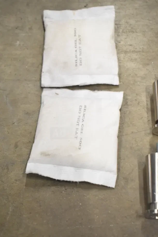 Two silica gel packets labeled "SILICA GEL 500 DO NOT EAT" on a surface, next to a metal part.