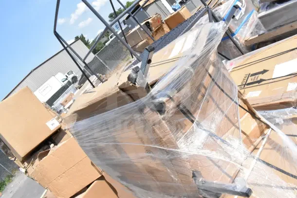 Stack of wrapped cardboard boxes containing brand new scratch and dent items including chairs, a work table, and a housekeeping cart.